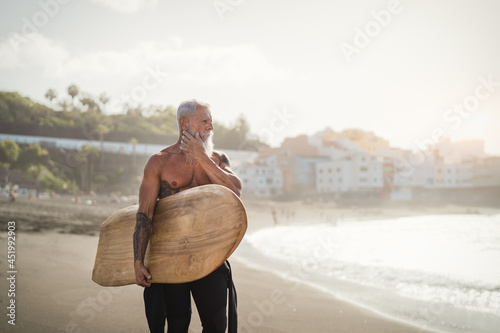 Senior male having fun surfing during sunset time - Fit retired man training with surfboard on the beach - Elderly healthy people lifestyle and extreme sport concept