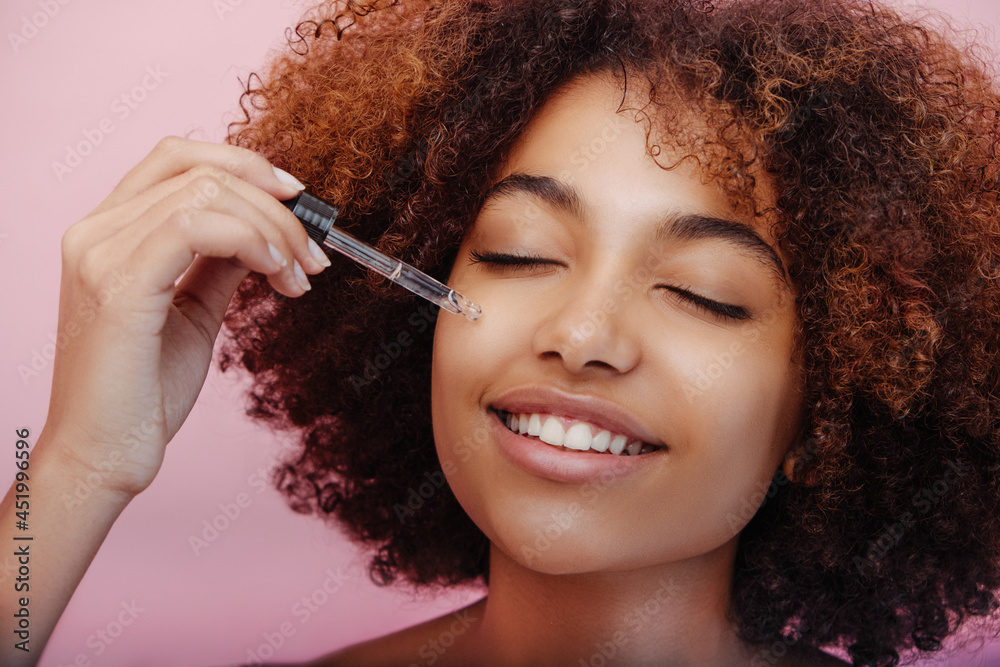 Obraz premium Mixed race young woman with short curly brown hair and perfect makeup holds pipette and touches face with closed eyes closeup