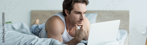 thoughtful man looking at laptop while lying in bed, banner