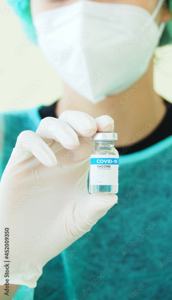 Vertical image Select focus Covid-19 vaccine bottle in hand with medical gloves. Blurred nurse wear mask as background. Urgent control Coronavirus outbreak situation. Health and Medical.