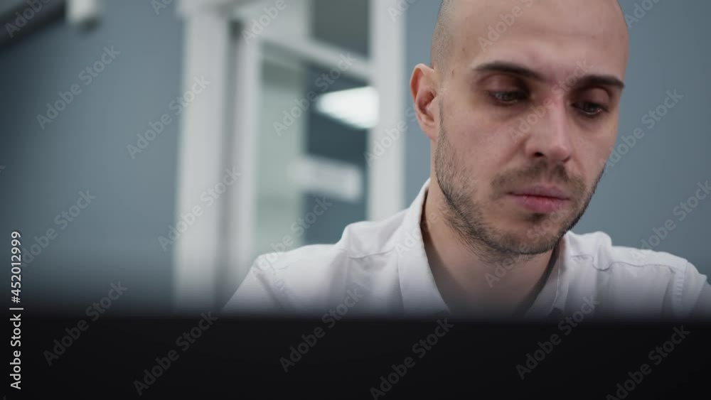Closeup of office man having problems, tired of working in front of the laptop. Young bald man sitting at business office, gimbal pan shot left to right
