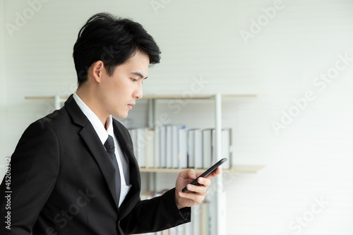 Portrait young businessman in black formal suit using mobile phone online meeting in modern office with business books shelf in the background. Young Asian man holding smartphone for business work.