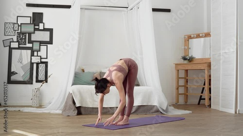 A slender, flexible, caucasian middle-aged woman with black hair in a beige tracksuit performs standing asanas tadasana on a purple yoga mat in a bright room with a bed, a mirror, a window photo