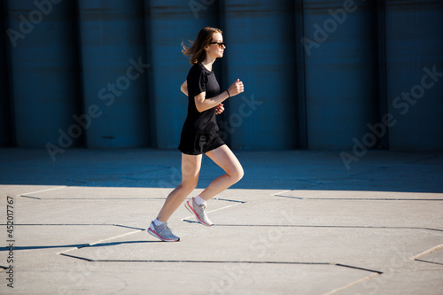 a white girl with short blonde hair in black clothes and light sneakers runs against the background of a gray concrete wall. photo motion drive