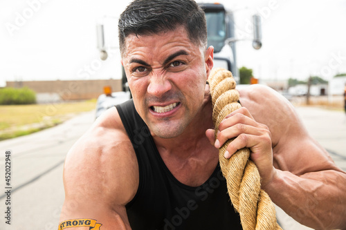 Muscular Hispanic man wearing tank top with Strong tattoo on his bicep, Straining to pull semi truck with large tow rope  photo