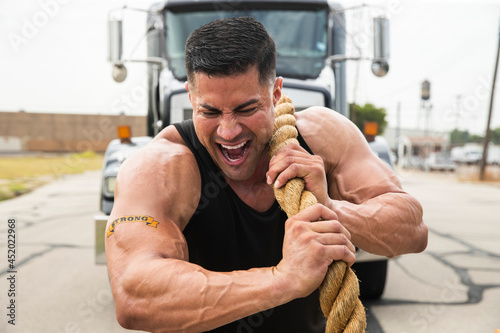 Muscular Hispanic man wearing tank top with Strong tattoo on his bicep, Straining to pull semi truck with large tow rope  photo