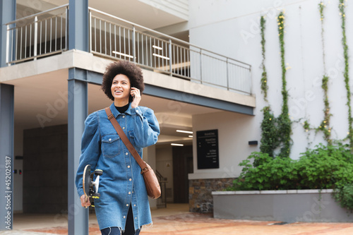 Smiling african american female business creative using smartphone and holding skateboard photo