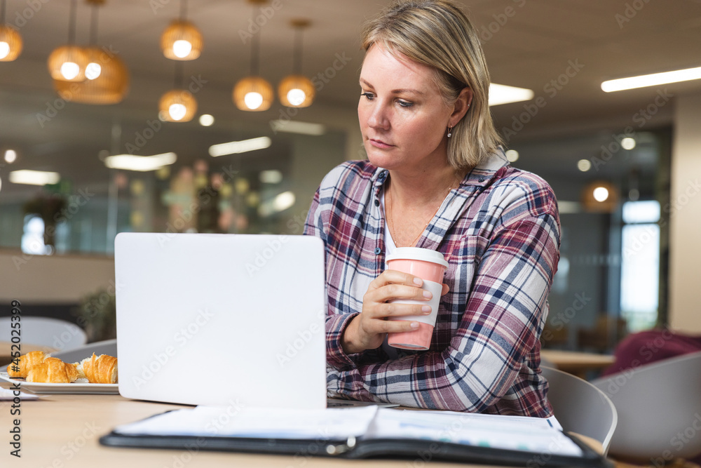 Thoughtful caucasian female business creative holding coffee and using laptop at workplace cafeteria Thoughtful caucasian female business creative holding coffee and using laptop at workplace cafeteria