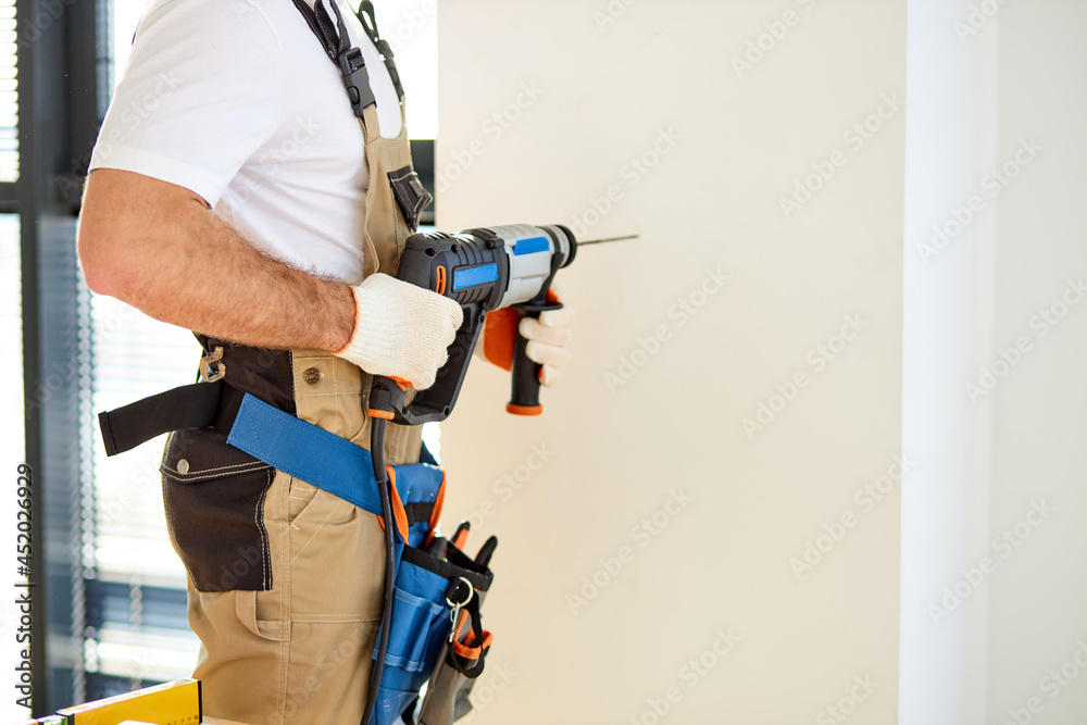 Cropped Professional Handyman In Overall Using Electric Drill For Making Hole In Wall, Handsome Guy Working With Power Tools, Home Renovation, Repair. Side View Cropped Professional Handyman In Overall Using Electric Drill For Making Hole In Wall, Handsome Guy Working With Power Tools, Home Renovation, Repair. Side View