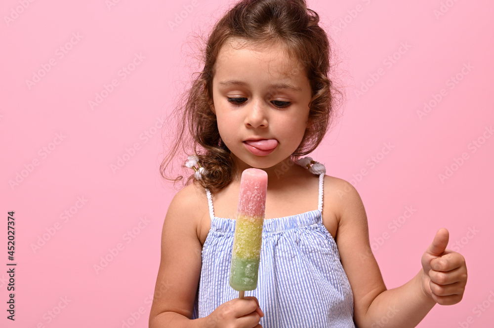 Headshot 4 years pretty baby girl with healthy vegan ice cream popsicle in hand and showing thumb up.Pink background, copy space . Summer dessert, cheerful summer mood concept Headshot 4 years pretty baby girl with healthy vegan ice cream popsicle in hand and showing thumb up.Pink background, copy space . Summer dessert, cheerful summer mood concept