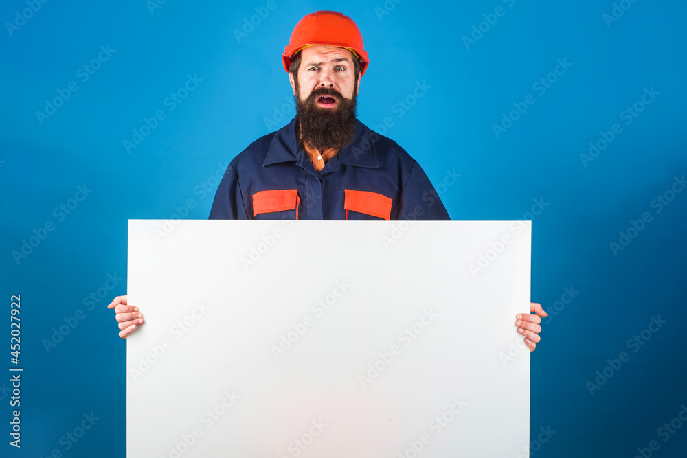 Confused builder with empty advertising banner. Bearded worker in protective clothing shows blank sign board ready for your text. Confused builder with empty advertising banner. Bearded worker in protective clothing shows blank sign board ready for your text.