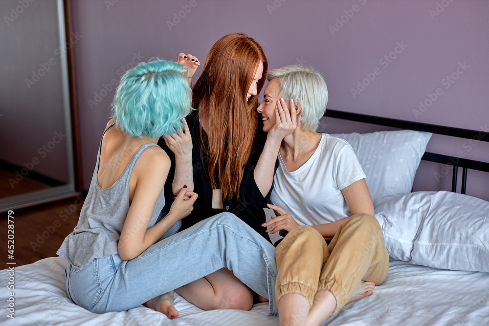 Threesome concept. Three awesome lesbian ladies in domestic clothes sit on  bed flirting, hugging, women in love, lesbian, same sex females enjoy  spending time together, going to have sex. side view Stock