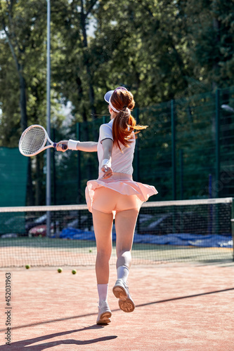 Young redhead woman in short skirt playing tennis. Female hits the ball with racket. Outdoor shot of slim slender fit lady in sportive wear. Youth, flexibility, power and energy. Rear view portrait