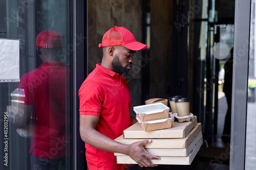 Food delivery service by afro american courier. Courier man in red uniform with pizza boxes, coffee cups, ready to serve clients customers. Confident african guy came to modern house, stands waiting