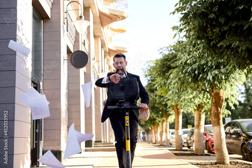 Man in formal suit Riding an Electric Scooter, hurry up, having no time. Modern Entrepreneur Uses Contemporary Ecological Transport to Go at Office Meeting, documents fly in different directions photo