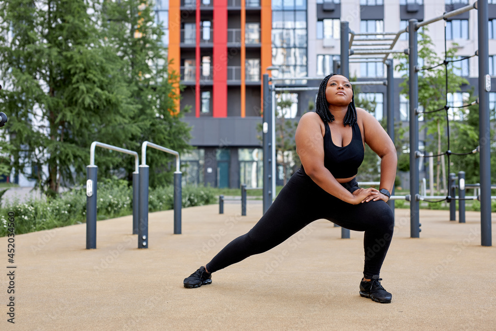 Fat afro american woman doing lunges in summer in city, training outdoors. Woman in black tracksuit sportswear leggings top sneakers, warms up muscles before workout. copy space, side view