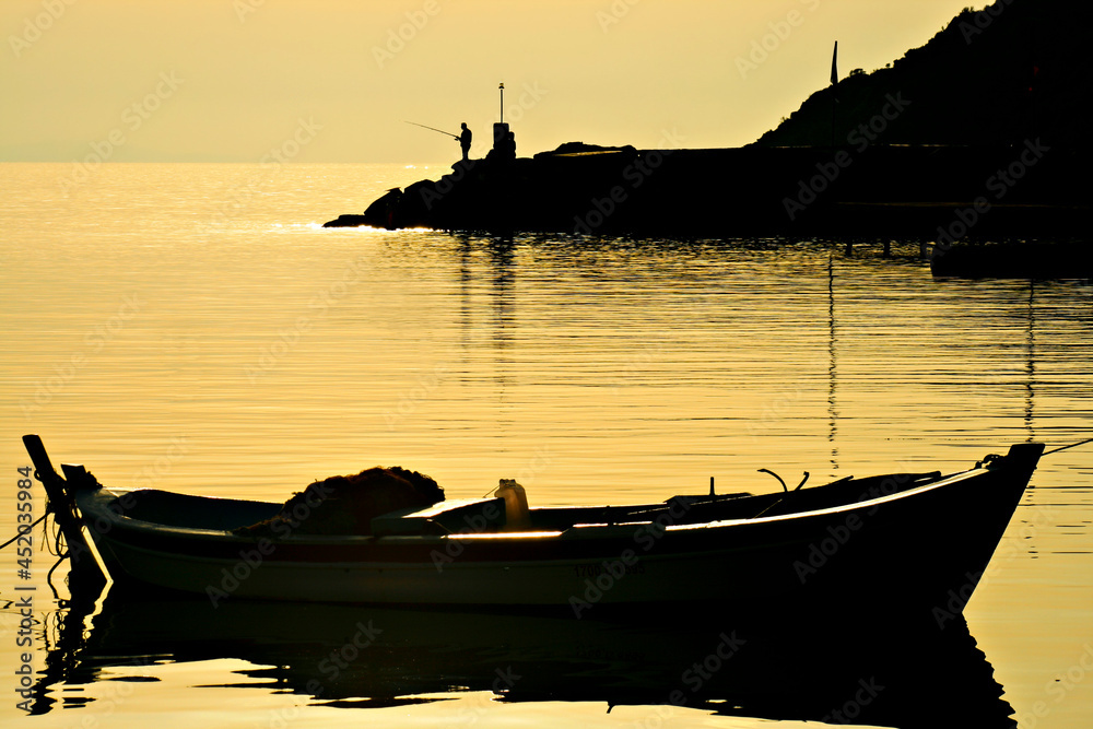 Fishing boat and fisherman in silhouette, at the sunset, along the Aegean Sea, in Assos, Turkey