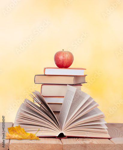 opened book with red apple on the stack of books and maple leaf on the yellow background 	