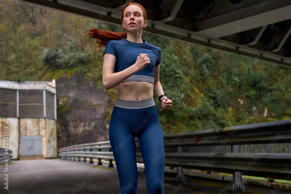 Portrait red-haired cute woman jogging on bridge alone in the morning, copy space. Young caucasian athlete woman exercising to lose weight. On modern bridge in countryside, urban sport concept