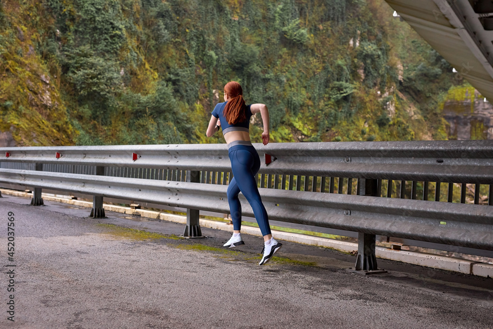 Rear view On Slim redhead Powerful lady jogging on modern bridge, view from back. urban sports, running in the morning, outdoors. copy space. sport, healthy lifestyle. sport and activity concept