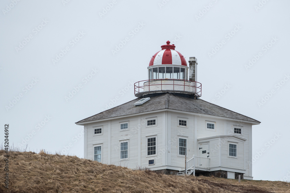 A historic square white and red wooden tower lighthouse with a round light room. There are a number of small windows on all sides of the building.  The background has lots of white fluffy clouds. 