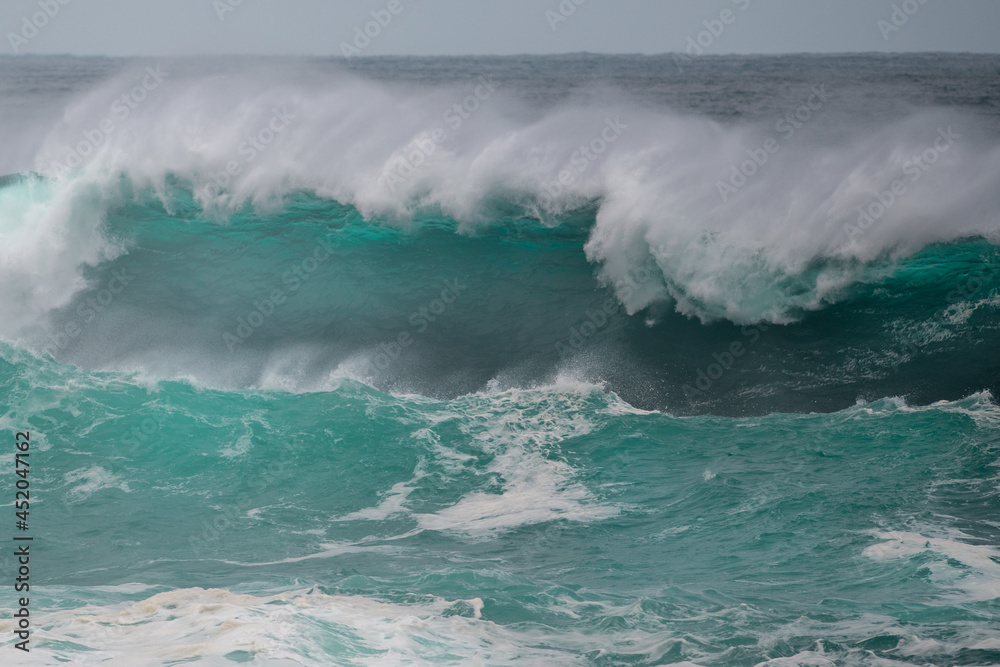 Obraz premium An angry turquoise green color massive rip curl of a wave as it barrels rolls along the ocean. The white mist and froth from the wave are foamy and fluffy. The ocean in the background is deep blue. 
