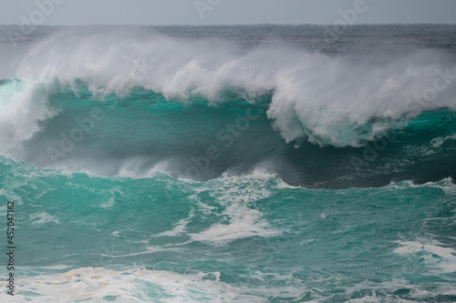 An angry turquoise green color massive rip curl of a wave as it barrels rolls along the ocean. The white mist and froth from the wave are foamy and fluffy. The ocean in the background is deep blue. 