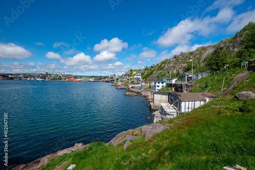 The hillside of St. John's Harbour, Newfoundland, on a sunny day, under blue sky and white clouds. The colorful wooden houses are scattered along the hillside with the blue ocean in the foreground. 