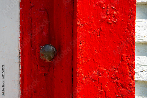 A closeup of an antique rusty iron decorative metal door handle on a brightly painted vibrant red wooden door. The vintage door is next to a white wall and glass window that's paint is peeling. 