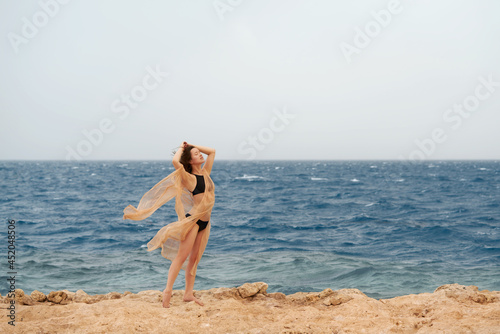 Pretty woman posing in the sea. Young caucasian woman in black swimsuit and golden dress sitting on stone shore