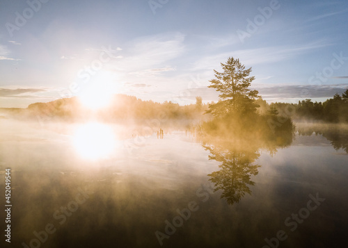 A sunrise view on the lake with a tree on the small island and the sun beams coming through the fog