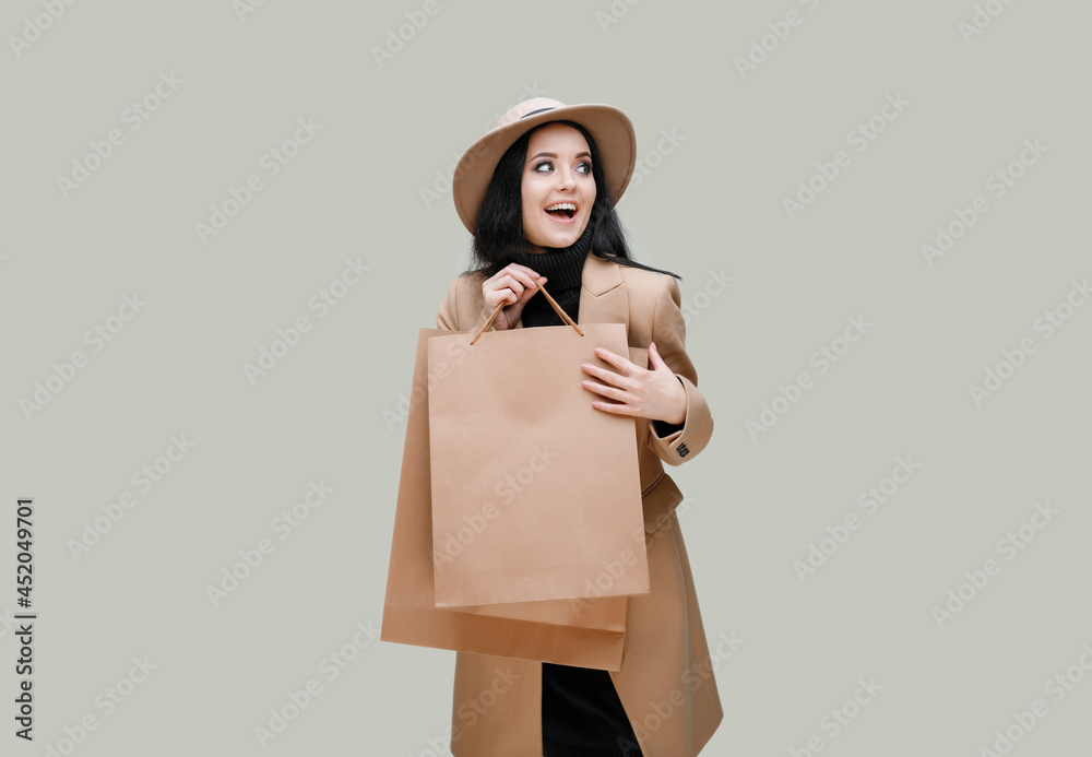 Portrait of charming lady in beige classic coat and hat with shopping bags in hands on gray background. Happy beautiful woman. Portrait of charming lady in beige classic coat and hat with shopping bags in hands on gray background. Happy beautiful woman.