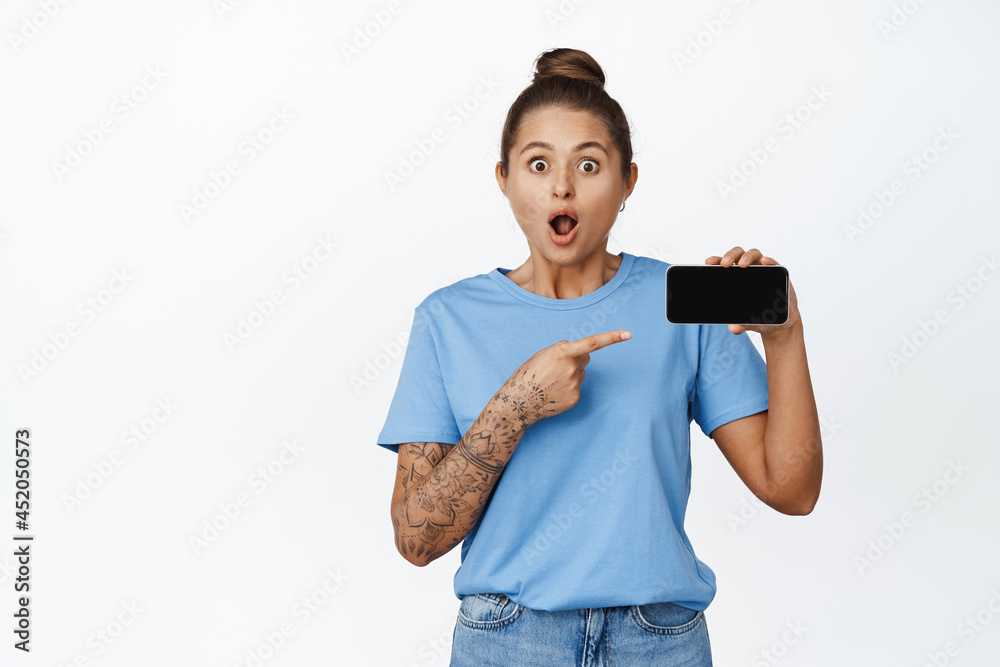 Portrait of girl looking amazed and showing mobile phone screen, pointing finger at smartphone, white background Portrait of girl looking amazed and showing mobile phone screen, pointing finger at smartphone, white background
