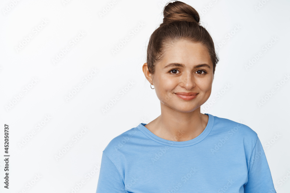 Close up portrait of young caucasian woman with hair combed in bun, smiling happy at camera, standing in blue tshirt against white background Close up portrait of young caucasian woman with hair combed in bun, smiling happy at camera, standing in blue tshirt against white background