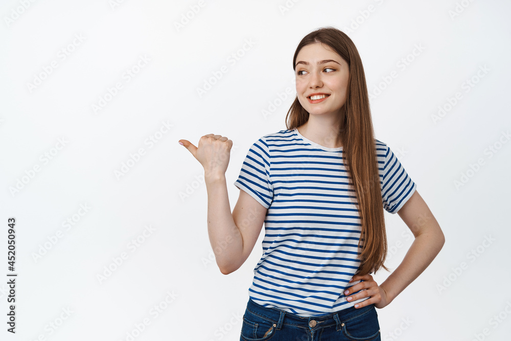 Young smiling woman, pointing and looking left with relaxed, happy face expression, standing in casual tshirt over white background Young smiling woman, pointing and looking left with relaxed, happy face expression, standing in casual tshirt over white background