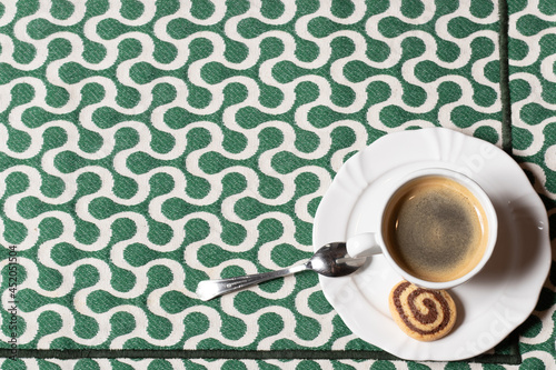 Hot black coffee, served on white crockery, with a small spoon and a two-color biscuit, served over a placemat with a white and green geometric print. With space for text.