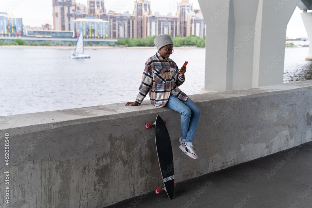 Black skater girl sit at concrete bridge with smartphone read message in email chat application outdoors. Happy woman with longboard scrolling web browser using wireless internet. Technology concept