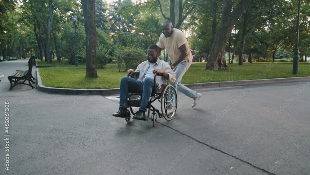 Mature adult black man walking in summer park with paralyzed non-mobile african guy, men having fun on weekend. Cheerful father rotates his son in wheelchair. Full life of disabled people concept