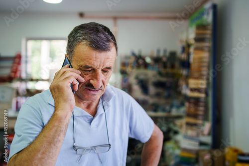 Senior caucasian man using mobile phone talking at work at the store in day - male entrepreneur with mustaches and eyeglasses talk about business real people copy space