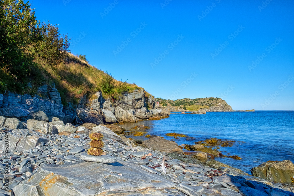 Rugged rock cliff with mountains in the background. The ocean is blue and calm at the land's edge. There are trees on the cliff and yellow grass sloping down. Sea urchins are stacked like a person.