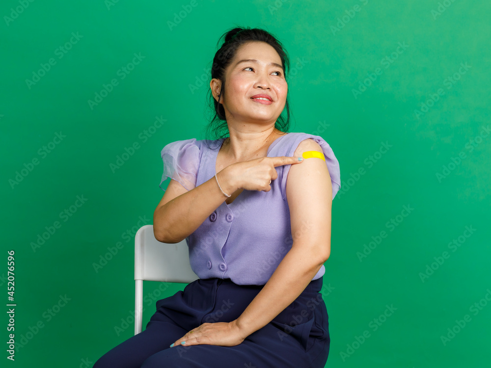Studio shot of Asian middle aged woman  sit smile pointing at yellow plaster bandage on arm after receive coronavirus covid 19 vaccination by doctor at clinic on green background