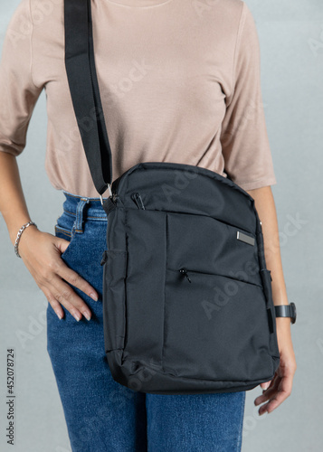 Closeup studio shot of female model in brown long sleeve shirt and jeans stand posing hanging fashionable trendy urban black multipurpose unisex handbag with long shoulder strap on grey background