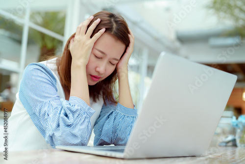 A beautiful Asian woman in a white shirt sits in front of a laptop computer with a stressed face clutching her head while working in a bakery.