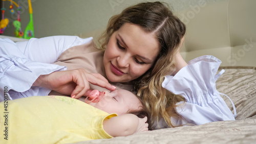 Joyful young mother with long loose hair lies on large bed near sleeping with pacifier in mouth baby girl in yellow bodysuit in bedroom closeup