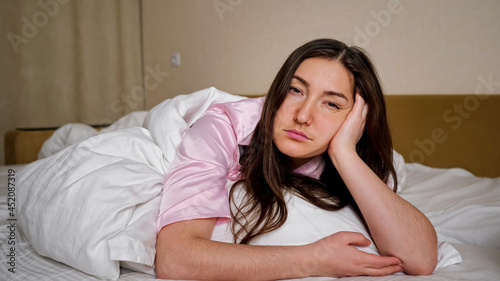 Sad unhappy dark haired woman in pink pajama sighs deeply lying lonely on king size bed with clean white linen in bedroom close view