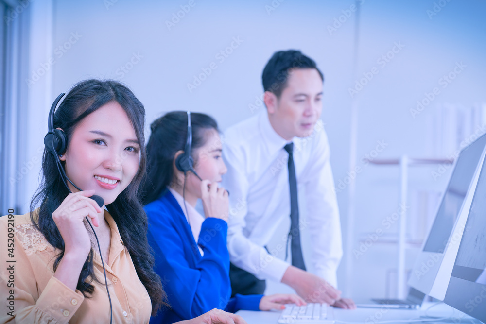 Asian call center female agent smile with headset and her team working on background in office for support customer Asian call center female agent smile with headset and her team working on background in office for support customer