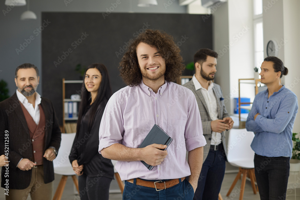 Portrait of happy businessman, team leader, business coach or motivational speaker. Young man with long curly hair standing in office in front of group of employees, holding notebook and smiling Portrait of happy businessman, team leader, business coach or motivational speaker. Young man with long curly hair standing in office in front of group of employees, holding notebook and smiling