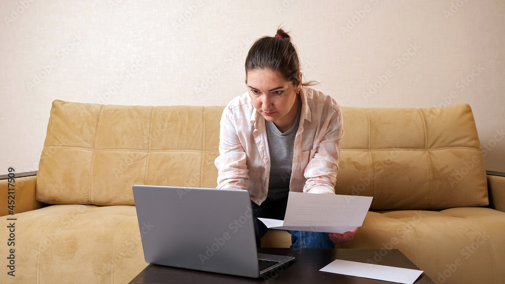 Pretty woman with bill paper calculates payments on modern laptop sitting on beige comfortable couch Pretty woman with bill paper calculates payments on modern laptop sitting on beige comfortable couch