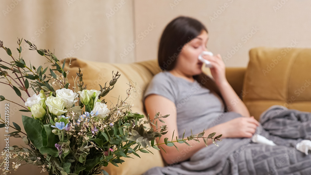 Blurred lady with flowers allergy sneezes in white paper napkin sitting on brown sofa with coloured bouquet on foreground at home close view Blurred lady with flowers allergy sneezes in white paper napkin sitting on brown sofa with coloured bouquet on foreground at home close view