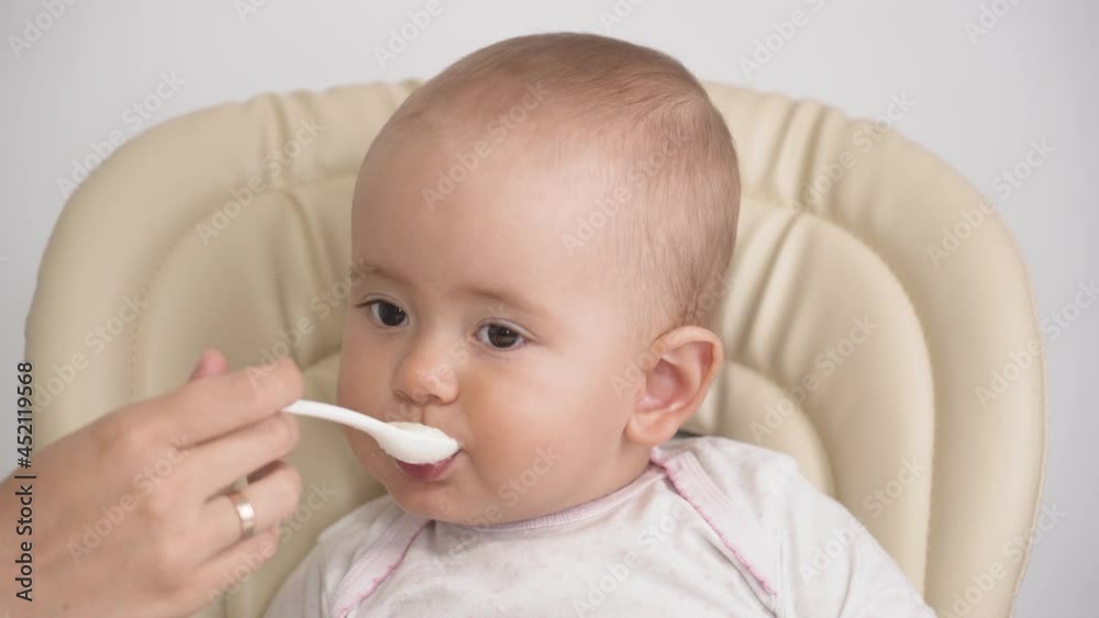 Mom feeds the baby with a spoon of oatmeal porridge. The child is sitting on a high chair and making faces. Close-up of the baby's portrait Mom feeds the baby with a spoon of oatmeal porridge. The child is sitting on a high chair and making faces. Close-up of the baby's portrait